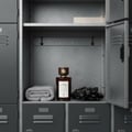 Maison Alhambra Smoked Oud & Vanilla by Maison Alhambra 100ml EDP — A dark glass perfume bottle with a wooden cap sits on a shelf inside a grey locker, next to a rolled grey towel and black gloves.