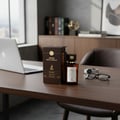 Maison Alhambra Smoked Oud & Vanilla by Maison Alhambra 100ml EDP — A brown box and a glass perfume bottle with a gold cap and label, placed on a wooden desk next to a laptop, watch, and glasses.