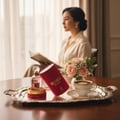 Maison Alhambra So Candid by Maison Alhambra 100 ml EDP — A woman reads a book by a window, with a tray of perfume, roses, and coffee in the foreground.