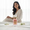 Maison Alhambra So Candid by Maison Alhambra 100 ml EDP — Pink perfume bottle with gold accents and a clear spherical cap, placed on a white bedspread with two small white flowers beside it. A woman in a white robe is blurred in the background.