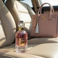 Maison Alhambra So Candid by Maison Alhambra 100 ml EDP — Pink perfume bottle with gold accents and a clear cap sits on a car seat next to a blush pink handbag with a tassel.