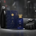 Maison Alhambra True Word by Maison Alhambra 100ml EDP — Blue perfume bottle with gold cap, navy blue box with gold design, black water bottle, and black gym glove on a grey surface.