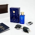 Maison Alhambra True Word by Maison Alhambra 100ml EDP — Blue perfume bottle with gold cap, matching blue box with gold swirls, sunglasses, passport, and car key fob on a white surface.