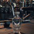 Maison Alhambra Victorioso by Maison Alhambra 100ml EDP Men — Clear glass perfume bottle with a textured silver cap, featuring a winged logo and text 'VICTORIOSO' and 'ACEONIDIA', placed on a gym floor with dumbbells and weight racks in the background.