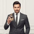 Maison Alhambra Victorioso Heroic by Maison Alhambra 100ml — Man in a grey suit holding a gold-accented brown perfume bottle with a white background.