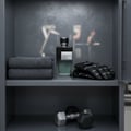 Maison Alhambra Yeah by Maison Alhambra 100ml EDP Men — A dark glass perfume bottle with a silver cap sits on a shelf next to folded grey towels and black workout gloves, with dumbbells visible on the shelf below.