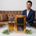 Maison Alhambra Your Touch Intense by Maison Alhambra 100ml — A clear glass perfume bottle with amber liquid and a silver ribbed cap, next to its gold and black box, with a man in a suit blurred in the background.
