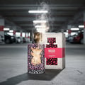 Mood Perfume Mood Pretty For Her 100 ml EDP — A perfume bottle with a pink and black abstract pattern and a gold starburst design, emitting smoke, stands next to its matching box in a dimly lit parking garage.