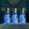 Osma Osma Ambry A Fresh & Fly 75ml EDP — Three blue glass perfume bottles with gold caps, each featuring a rose emblem and text labels, arranged on a textured surface against a dark, patterned backdrop.