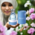 Osma Cotton C Rose & Sandalwood 75ml — Perfume bottle and cylindrical box with floral motifs, a piece of sandalwood, and a blurred woman in a hijab in the background with pink flowers.