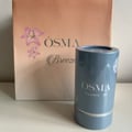 Osma Flowers W Osma Sandalwood 75ml — A blue cylindrical container with 'OSMA Flowers' W' text sits in front of a peach-colored paper bag with pink flowers and 'OSMA Breeze' text.