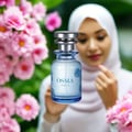 Osma Fruity O Mandarin & Amber 75ml EDP — A clear glass perfume bottle with a silver cap, featuring blue floral designs, is placed in front of a blurred background of pink flowers and a woman wearing a white hijab.