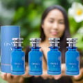 Osma Osma O Tobacco & Pear 150ml EDP — Four blue perfume bottles with silver caps, one tall blue box, and a blurred woman in the background with greenery and a white flower.