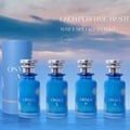 Osma Osma A Tropical & Soft 150ml EDP — Four blue perfume bottles with silver caps and floral designs, lined up on a reflective surface with a blue sky and clouds background. A cylindrical box is to the left.