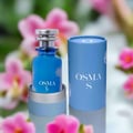 Osma Osma S – Pepper & Sandalwood EDP 150 ml — Blue perfume bottle with silver cap and floral motif, standing next to its blue cylindrical box, with a soft focus background of pink flowers.