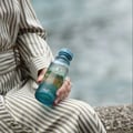 Osma Osma Portofino Summer Purity 150ml EDP — Woman in striped dress holding a blue glass perfume bottle with a scenic label, outdoors by water.