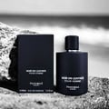 Pendora Noir En Leather Pour Homme — Black perfume bottle and box displayed on sand and rocks, with a blurred beach background.
