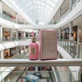 Paris Corner Qissa Pink by Paris Corner 100ml for Women — Pink fluted perfume bottle with gold cap and label, next to its matching pink and gold patterned box, displayed on a silver railing in a shopping mall.