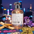Paris Corner Rich Santal Emir by Paris Corner — A clear glass perfume bottle with a silver cap, filled with amber liquid, sits on a dark surface with neon lights. Palo santo sticks, cardamom pods, and purple flowers are arranged around it.