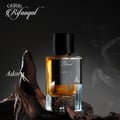 Paris Corner Rifaaqat Adorn by Paris Corner 85ml EDP — Amber liquid in a clear glass bottle with a black cap, resting on dark wood with wisps of smoke in the background.