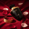 Rue Broca Carino Black Pour Femme 100ml EDP — Black perfume bottle with gold accents, resting on red satin fabric, surrounded by scattered rose petals and a gold cuff bracelet.