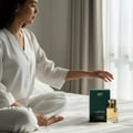 Rue Broca Luminus Pour Femme 100ml EDP — Woman in white pajamas sitting on a bed, reaching towards a green perfume box and gold-capped bottle.