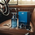 Rue Broca Luminus Pour Homme — Botol minyak wangi kaca biru dan kotak sepadan bersama cermin mata hitam dan kunci kereta di atas tempat duduk kereta kulit coklat.