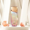 Rue Broca Nexa Musee by Rue Broca – 100 ml for Women, a shimmering iridescent bottle with a gold cap, displayed on a white pedestal with geometric peach-colored props and spheres.