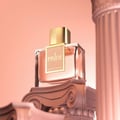 Rue Broca Pride Pour Femme — A clear glass perfume bottle with a gold cap and a beige label that says "PRIDE POUR FEMME", displayed on an ornate, carved pedestal against a soft pink background.