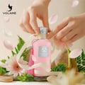 Volare Darlene Pour Femme – Hands tying a ribbon around a pink perfume bottle, surrounded by flowers, petals, and stones.