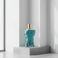 Volare Le Beau Gladiator Twilight Volare 100ml for Man — A clear glass perfume bottle shaped like a muscular male torso with blue liquid, topped with a gold cap, displayed on a grey stone pedestal against a white background with soft shadows.