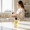 Volare Lustre Amour – Botol minyak wangi kaca jernih bertutup hitam terletak di atas permukaan marmar putih, dengan seorang wanita sedang menuang cecair di latar belakang.