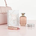 Volare Amber Lychee Kenzie by Volare – 100 ml for Her perfume bottle, lipstick, compact powder, and gift box arranged on a pink surface with a pink handbag in the background.