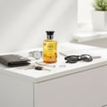 Volare Rise & Shine by Volare – 100 ml Unisex, amber liquid in a clear glass bottle with a black cap, placed on a white surface with a wallet, watch, keys, sunglasses, and a notebook.