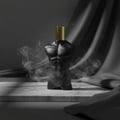 Volare Elixir Gladiator Twilight by Volare 100ml Man — Black torso-shaped perfume bottle with gold cap, surrounded by wisps of smoke, on a marble surface with draped dark fabric.