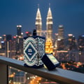 Zimaya Happy Oud — Botol minyak wangi biru gelap dengan corak geometri emas yang rumit dan tulisan "HAPPY OUD" dipamerkan di atas pagar balkoni pada waktu malam, dengan latar belakang langit Kuala Lumpur yang diterangi lampu dan Menara Berkembar Petronas. Kotak kecil berwarna emas yang terbuka diletakkan di sebelah