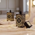 Zimaya Happy Oud by Zimaya 100ml Extrait Unisex — Dua botol minyak wangi hitam dengan rekaan art deco emas dan hijau di atas permukaan marmar bersama kelopak mawar putih bertaburan. Penutup hitam kecil beraksen emas terbaring di sisinya.