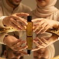 Zimaya Ilham Al Oud by Zimaya 100ml EDP Unisex — Botol minyak wangi kaca berlabel emas dipegang tangan di atas permukaan cermin, memantulkan botol dan tangan.