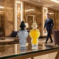 Zimaya Infrad Noir by Zimaya 100ml EDP untuk Lelaki — Dua botol pewangi berhias, satu bertutup hitam bercecair kelabu, satu lagi bertutup emas bercecair kuning, dipamerkan di atas meja kaca di lobi hotel mewah dengan seorang lelaki berbaju sut berjalan di latar belakang.