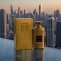 Zimaya Noor Oud Dubai 100ml EDP Unisex — Gold perfume bottle and box set against a blurred city skyline at sunset.