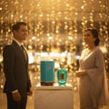 Zimaya Rabab Blue by Zimaya 100ml EDP Unisex — A man in a suit and a woman in a dress stand smiling on either side of a teal perfume bottle and its box, displayed on a marble pedestal against a bokeh of golden lights.