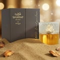Zimaya Sharaf Divine By Zimaya 100 ml Extrait Unisex — Brown textured box with gold Arabic and English text, next to a clear glass perfume bottle with a textured white cap, all resting on a bed of fine sand with wood pieces.