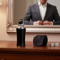 Zimaya Taraf Black by Zimaya 100ml EDP Men — Botol minyak wangi hitam dengan perincian perak, tali leher hitam yang digulung, dan butang lengan baju perak di atas meja kayu, dengan pantulan lelaki bersuit di cermin di belakang.