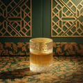 Zimaya Sharaf Divine By Zimaya 100 ml Extrait Unisex — A clear glass perfume bottle with gold liquid sits on a green and gold patterned fabric, with a decorative gold lattice background.