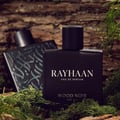 Rayhaan Woody Noir Eau De Parfum — Two black perfume bottles, one in focus with "RAYHAAN" and "WOOD NOIR" text, surrounded by moss and tree bark. | Dua botol minyak wangi hitam, satu dalam fokus dengan teks "RAYHAAN" dan "WOOD NOIR", dikelilingi oleh lumut dan kulit kayu.