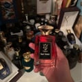 Bujairami Red Static by Bujairami Eau de Parfum 100ml — Hand holding a red glass perfume bottle with a gold cap and black label, surrounded by other perfume bottles and display items.