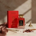 Bujairami Red Static by Bujairami Eau de Parfum 100ml — Red perfume bottle and matching box on a marble surface with silk fabric, dried rose petals, and a cinnamon stick.