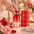 Osma Romance by Osma Perfumes 150ml EDP Women — A hand with a diamond ring reaches for a red perfume bottle, with a white handbag, roses, and candles in the background.