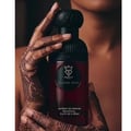 Bujairami Sacred Skin 100ml Extrait Unisex — Close-up of hands with intricate henna designs holding a dark red perfume bottle with a black cap and a circular logo.