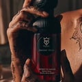 Bujairami Sacred Skin 100ml Extrait Unisex — Close-up of hands with intricate henna designs holding a dark red perfume bottle with a black ribbed cap.